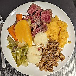 A white plate holds a meal of sliced roast beef, mashed sweet potatoes, stuffing, boiled potatoes, carrots, and cabbage, set against a dark, textured surface.